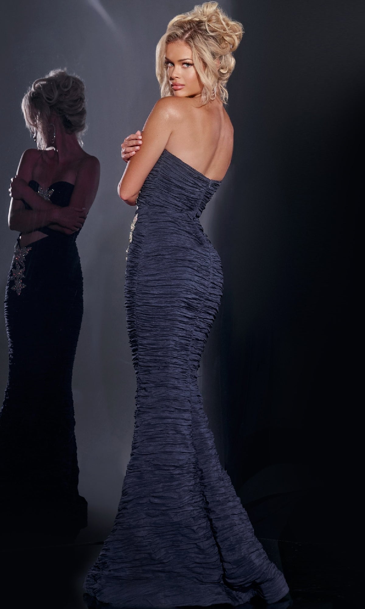 A woman with blonde hair stands with arms crossed in the elegant, textured strapless Jovani 40174 long prom dress, looking over her shoulder as her reflection appears in the background.